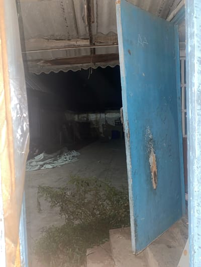 The image shows an open, weathered blue metal door leading into a dimly lit, seemingly abandoned or under-construction structure. The door is marked with white chalk writing that appears to be "AA" and other illegible marks. To the left of the door, the entrance is partially obscured by a transparent plastic sheeting. Inside the structure, the floor is concrete, and there are various objects scattered around, including what appear to be large, light-colored tarps or fabric sheets. Some debris and what looks like a blue barrel are visible further inside. The ceiling appears to be constructed of corrugated metal and wooden beams, showing signs of disrepair. A small bush is growing near the entrance on the right side of the image. The scene suggests an outdoor or semi-outdoor space, possibly a yard or a makeshift shelter, located in Кедей-Арык, Kyrgyzstan.