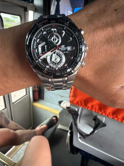 The image shows a close-up of a person's wrist wearing a black and silver digital and analog watch. The watch face displays multiple subdials and time markers. The background is out of focus, but appears to be an indoor environment, possibly a vehicle or a public transport setting, with some seats and windows visible. The person's arm and hand are visible, with some tattoos on the arm. The lighting suggests it is daytime. There is no discernible connection to Bethamcherla, India in the image.