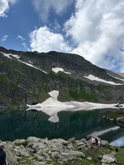 The image shows a serene mountain lake scene in Panichishte, Bulgaria. The lake's dark, reflective water mirrors the imposing, rocky mountain range that rises dramatically behind it. Patches of snow cling to the mountainside, contrasting with the green vegetation. The sky above is a mix of blue and scattered white clouds, suggesting a pleasant day. In the foreground, a group of four individuals, two adults and two children, are gathered on a rocky shore, some appearing to be observing the view. One person, possibly a young woman, is seated on a rock with their back to the camera, wearing a black top. Another person is visible on the far left, also with their back to the camera, wearing a black top. A black backpack rests on the ground near the group. The overall impression is one of natural beauty and peaceful outdoor activity.