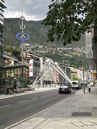 The image captures an urban street scene in Andorra la Vella, set against a backdrop of steep, densely forested mountains under an overcast sky. In the foreground, green leaves from a tree partially frame the left side of the shot.
A wide, grey pavement on the right leads to a two-lane asphalt road with white markings. A black sedan and a white commercial van are parked or driving on the road. A pedestrian, wearing a light shirt and dark trousers, stands on the sidewalk next to the white van.
A prominent feature in the midground is a large, distinctive white architectural structure, resembling a fan-like archway or support system, with radiating struts and spherical joints. This structure is part of a modern building complex. Below it, outdoor seating areas with white tables and chairs are visible, with a few people seated.
Rising vertically from the street is a tall sign bearing large, stylized letters. The silver letters "CALDEA" are clearly visible, along with a prominent blue "A" which is part of the "Caldea Aqua" logo, indicating the famous spa complex.
The background is dominated by towering, green mountains, their slopes covered in trees, with scattered buildings and infrastructure blending into the natural landscape. The overall setting is a modern city integrated into a mountainous environment.
Visible text includes "CALDEA" and the blue "A" from the Caldea logo, a blue "P" parking sign, "OPEN" on a shop window, and "PLAN B" on a building near the white archway. Other smaller signs and digital advertisements are also present but largely unreadable due to distance. The time of day appears to be daytime, with soft, diffused light suggesting a cloudy weather condition.