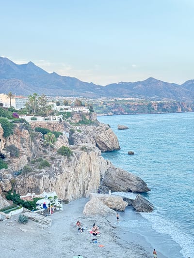 This image depicts a view of the coastline near the Balcón de Europa in Nerja, Spain, characterized by its dramatic cliffs, azure waters, and a small beach area. In the foreground, a sandy beach is populated by several individuals relaxing or wading in the shallow water. A white building with a red-tiled roof, likely a small restaurant or chiringuito, is nestled at the base of the cliffs, adorned with white awnings and greenery. The cliffs themselves are rugged and textured, with sparse vegetation clinging to their surfaces. Several large rocks and boulders are scattered along the shoreline and in the water.

Further inland, the landscape rises to reveal a town built on the clifftops, featuring white buildings and hotels, many with balconies overlooking the sea. Palm trees and other Mediterranean flora dot the landscape. In the background, a mountain range stretches across the horizon, its peaks softened by the hazy, pale blue sky. The sea is calm and a vibrant turquoise, with gentle waves lapping at the shore. The overall impression is one of a serene coastal scene in a Mediterranean town.