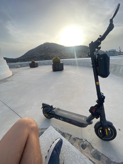An outdoor, eye-level, wide shot captures a black electric scooter parked on a concrete surface with a mountain range and a harbor in the background. The sun, partially obscured by clouds, shines brightly in the upper right portion of the frame. The scooter is positioned in the right foreground, angled towards the viewer, with its handlebars reaching towards the top of the image. A black bag is attached to the handlebars. In the left foreground, a person's bare legs and a dark blue and white sneaker are visible, suggesting they are seated on a concrete ledge. Two large, round cacti in dark planters are spaced out on the concrete surface behind the scooter. The concrete area appears to be a promenade or rooftop, with a white structure on the far left and a white barrier separating it from a body of water where ships and cranes are visible in the distance, indicating a harbor setting. The overall scene suggests a coastal location in Cartagena, Spain, likely a scenic viewpoint overlooking the harbor and city.