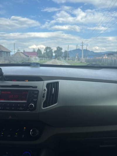 This is a photograph taken from inside a car, looking out through the windshield. The car's dashboard is visible in the foreground, with a radio display showing "88.10" and "18:18" on the right. The exterior scene depicts a rural landscape with houses and trees, a dirt road, and mountains in the distance under a cloudy sky. The time appears to be late afternoon. There are no people visible in the image. The image appears to be taken in Yabogan, Russia, based on the context.