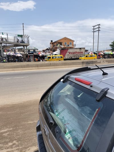 The image shows a street scene in Ibafo, Nigeria. A silver Kia car is in the foreground, partially obscuring the view. In the background, there's a bustling market area along a highway, with various stalls covered by umbrellas. Two yellow minibuses are visible, along with a large "Xtratherm" truck. An overhead pedestrian bridge spans the highway. There are people on the bridge and in the market area. Power lines stretch across the sky, and a few buildings are also visible in the background. The sky is partly cloudy. There is writing on the concrete barrier of the highway.