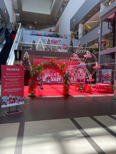 This media file captures an indoor shopping mall, likely located in Luanda, Angola, decorated for a festive occasion. The image is taken from an elevated perspective, looking down at the ground floor where a large Christmas-themed display is set up on a bright red carpet.
The central part of the display features a decorative white Ferris wheel with a Santa Claus cutout in front. A large arch made of red, green, and gold balloons, adorned with candy cane motifs and golden starbursts, frames a background featuring white city silhouette cutouts. Several Christmas trees, some with white "snow" flocking, are positioned around the display. Small red and green tables and chairs are also arranged on the red carpet.
On the left side of the frame, a large nutcracker figure stands next to an escalator. A few people are visible on the escalator, moving between floors. The setting suggests a daytime scene, with natural light entering from above, casting distinct diagonal shadows across the tiled floor on the right side of the image.
A prominent red sign titled "REGRAS" (Rules) lists several regulations in Portuguese: "NÃO TOCAR NOS ARTIGOS" (DO NOT TOUCH THE ARTICLES), "NÃO TRAZER ALIMENTOS E BEBIDAS" (DO NOT BRING FOOD AND DRINKS), "NÃO SE COLOCAR DEBAIXO DA RODA GIGANTE" (DO NOT GO UNDER THE FERRIS WHEEL), "NÃO CORRER PELO CENARIO" (DO NOT RUN AROUND THE SCENE), "NÃO USAR OBJETOS CORTANTES" (DO NOT USE SHARP OBJECTS). It concludes by encouraging visitors to "APROVEITE E GUARDE O MOMENTO TIRANDO FOTOS E MARQUE NOS STORIES @shoppingfortaleza.oficial" (ENJOY AND SAVE THE MOMENT TAKING PHOTOS AND TAG US IN STORIES @shoppingfortaleza.oficial). The overall atmosphere is festive and inviting, designed for family enjoyment and photo opportunities.