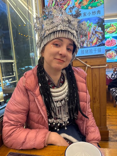 A Caucasian female is seated at a light wooden table inside a restaurant in Fenghuang, China, smiling at the viewer. She wears a large, ornate silver-colored Miao ethnic headpiece with leaf-like designs, bells, and subtle blue/green accents, over her dark braided hair. Her attire includes a salmon-pink padded puffer jacket, partially obscuring a patterned dark red, black, and white traditional top. A multi-layered silver-colored Miao necklace or collar, featuring intricate patterns, chains, and dangling bells, adorns her neck and chest. She has light pink eyeshadow and two small star-shaped glitter pieces on her right cheek. A white ceramic bowl with a blue rim rests on the table in the lower right foreground. The background behind her left side features a window revealing an out-of-focus evening city scene with bokeh lights. To her right, two brightly lit digital menu boards display colorful images of Chinese dishes, including "农家小炒肉" (Farmer's Stir-fried Pork) and "腊猪脸干锅" (Preserved Pork Face Dry Pot). Wooden wall paneling and a vertical wooden beam are visible within the restaurant interior.