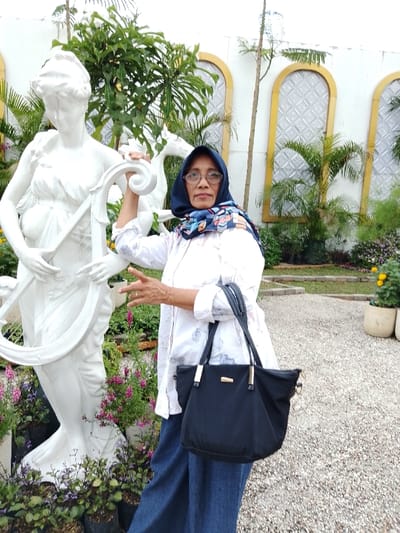 The scene is an outdoor landscaped area in Ngadirejo, Indonesia, featuring light-colored gravel ground. An adult female, wearing eyeglasses, a dark blue patterned hijab, a white patterned long-sleeved shirt, and dark blue wide-leg pants, stands to the right of a large white classical statue. The woman carries a dark blue handbag with black straps and gold-colored accents on her left shoulder. Her right hand grasps the statue's left arm, while her left hand is raised with fingers slightly spread, displaying a ring on her left ring finger. She gazes directly forward.
The primary statue depicts a draped female figure holding a lyre. Partially visible behind it is a second white statue, appearing to be a horse. Numerous green plants and flowering vegetation, including purple, pink, and yellow blooms, are present in the foreground and midground, some housed in black polybags and white planters. The background consists of a white building facade with three yellow-trimmed arched recesses, each fitted with patterned translucent panels. Various taller plants and slender trees are also scattered within the background vegetation.