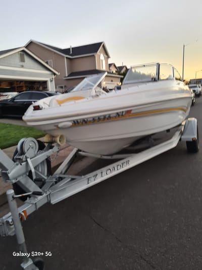 A white and orange boat is on a silver trailer. The boat is a Bayliner, model number is partially visible as "WN-434-NL". The trailer has "EZ LOADER" written on the side. The trailer is parked on an asphalt street, with a house and garage in the background. A street light is visible in the upper right corner. The sky is a pale orange, indicating either sunrise or sunset. The image is watermarked "Galaxy S20+ 5G".