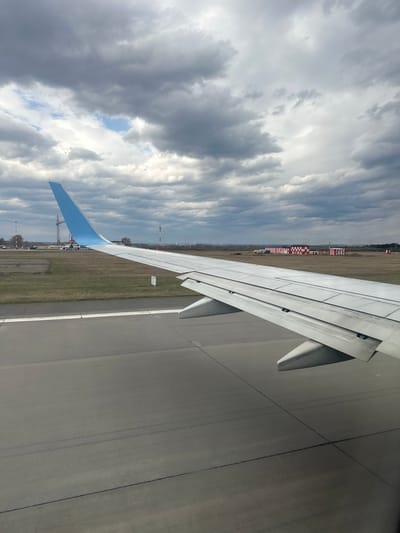 This media file displays a view from an airplane window, showing an aircraft wing on the ground at an airport. The image is oriented vertically.
On the left side of the frame, a cloudy sky dominates, featuring dense grey and white clouds with visible patches of brighter blue sky breaking through. The airplane's wing, predominantly white with a light blue wingtip, extends diagonally across the middle of the image, from the lower-left towards the upper-right. The wing shows mechanical details and panels.
To the right of the wing, a paved surface, likely an airport runway or taxiway, is visible, marked with subtle lines. Below the wing and beyond the paved area, a strip of dry, light brown grass separates the foreground from the background elements. In the distance, various airport structures can be seen, including distinctive red and white checkered markers, some smaller buildings, and a line of trees under the overcast sky.
The scene depicts an airplane on the ground, possibly taxiing or waiting for takeoff/landing. The time of day appears to be daytime, with an overcast sky. There is no discernible text visible in the image. The location is contextualized as Bolshoye Savino, Russia.