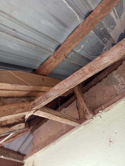 The image shows the underside of a corrugated metal roof supported by rough-hewn wooden beams. The wooden structure appears aged and somewhat deteriorated, with some beams showing cracks or exposed grain. Dust and debris are visible on the metal sheeting and wooden supports. The scene is consistent with the roof structure of a building in Gindin Dutse, Nigeria, suggesting a rural or less developed urban setting where such construction materials and methods are common. No people are present in the image. The overall impression is one of a functional but rustic building interior.