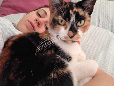 A young woman lies on a bed with blue and white striped bedding. She is wearing a light-colored top and has a pink pillow behind her. She looks tired and is gazing at the camera. A calico cat is lying on her chest, also looking at the camera. The cat has a white chest and paws, black and brown fur on its back, and a white face with black and brown markings. The cat's eyes are green. The location of the image is not in Birmingham, United Kingdom. The image was taken indoors.