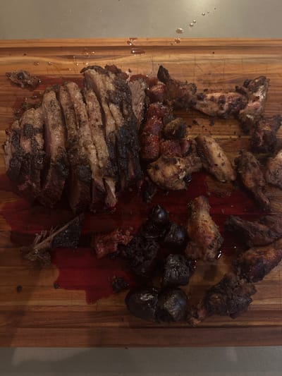 The scene depicts a wooden cutting board on a light-colored countertop in Miami Shores, United States of America. Dominating the left side of the board is a large cut of dark-crusted, sliced meat, likely beef brisket, showing internal pinkish-brown meat and fat marbling, with approximately eight visible slices. Red meat juices heavily pool beneath this meat and spread across the board. To the right, numerous cooked chicken pieces, primarily wings and drumettes, are scattered, exhibiting browned and lightly charred surfaces. Interspersed among the chicken are several smaller, reddish, sausage-like pieces and multiple dark, heavily charred, amorphous objects, possibly vegetables or dense meat scraps. A small bone fragment is visible in the lower-left section, partially submerged in the red liquid. No people are visible; the scene suggests post-preparation and pre-consumption of a meal.