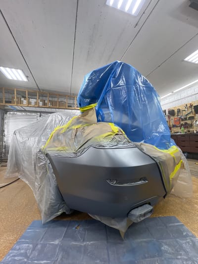 The image shows a car in a workshop, likely undergoing repair or painting. The rear bumper and parts of the car's body are covered in protective blue plastic sheeting and masking tape. The workshop environment features fluorescent lighting on the ceiling, shelves with boxes in the background, and tools mounted on a wall. The floor appears to be covered in a patterned material and a protective blue plastic sheet is laid out in front of the car. There are no people visible in the image. The location is identified as Veteran-2, Russia.