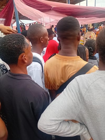 This image captures a crowded gathering in Ogbogbo, Nigeria, viewed from the perspective of an individual standing within the crowd. An overhead canopy, constructed from draped light pink and dark maroon fabric supported by blue metal poles, shelters the scene. In the foreground, multiple individuals are observed primarily from behind. A person on the far left wears a black short-sleeved polo shirt with white horizontal stripes on the collar, their head slightly turned to the right. Next to them, a person in a white long-sleeved shirt has a black strap, consistent with a backpack, over their right shoulder. To their right, a person in a mustard yellow ribbed t-shirt has a diagonal black strap across their back, extending from the left shoulder towards the right hip. Another individual, partially visible on the far right, wears a grey long-sleeved shirt with their left arm bent, hand resting on their hip. In the midground, additional attendees are visible, including one wearing a light-colored head covering patterned with Mickey Mouse figures. The background shows a dense crowd of people, some wearing blue, yellow, and green clothing, suggesting a communal event or assembly. Natural daylight illuminates the scene.