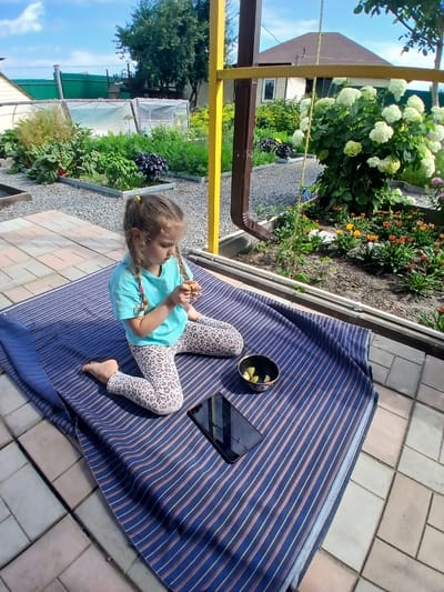In Maslova Pristan', Russia, a young girl with braided blonde hair sits on a striped blanket outdoors. She is wearing a turquoise t-shirt and leopard-print leggings. In front of her on the blanket are a small bowl with what appears to be lemon slices and a black tablet. The girl is holding an object in her hands, seemingly playing with it. The scene is set in a yard with a paved patio and a garden in the background, featuring various plants and flowers. A house and a greenhouse are also visible. The overall environment suggests a sunny day.