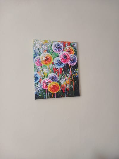 A colorful, abstract painting of dandelions is mounted on a light-colored, textured wall. The painting depicts several dandelions in various stages of bloom, with vibrant colors such as pink, orange, yellow, blue, and white. The background is a mix of green and white, suggesting a field of flowers or grass. The painting is rectangular and hangs vertically. The wall appears to be an interior wall. This scene is located in the city of Mamaivtsi, Ukraine.