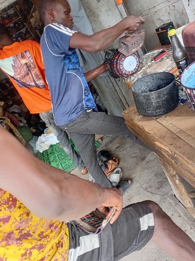 The image depicts an interior scene within the city of Sharada, Nigeria, likely a small workshop or market stall. In the center, a dark-skinned male wearing a blue short-sleeved athletic shirt with white stripes and blue camouflage patterns, and dark trousers, is standing and leaning forward. He is actively using a charcoal iron to press a round, multicolored traditional cap, which he holds steady with his left hand. His gaze is directed downwards at the cap. To the left, a second dark-skinned male, seen from behind, wears an orange short-sleeved shirt with a building graphic. In the immediate foreground, the right arm and upper leg of a third dark-skinned individual are visible; they wear a yellow and brown camouflage-patterned shirt and dark shorts with white side stripes, and hold a small, colorful woven item in their right hand.
On a rustic wooden table to the right, a black, worn metal pot, a partially visible blue woven basket, a brown glass bottle with a black label and yellow cap, and a black box with a green cap are arranged. The floor is concrete, displaying various items including a green patterned mat, discarded clothing, and two pairs of sandals (one brown leather, one black slide). The background is cluttered with shelves and miscellaneous objects, indicating a working environment.