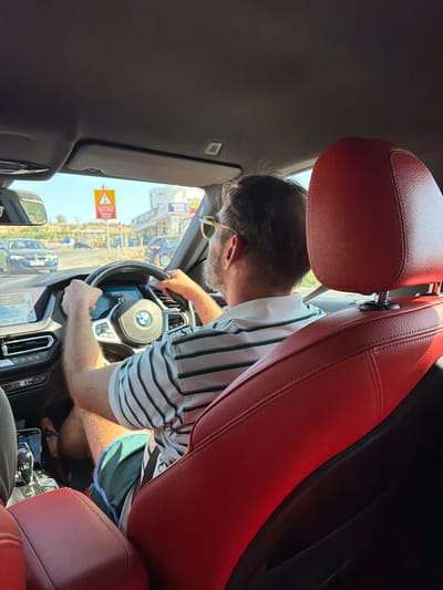 The image captures the interior of a car, looking forward from the passenger seat. A man with sunglasses and a striped polo shirt is driving, his hands on the steering wheel. He is wearing green shorts. The car's interior features red leather seats and a modern dashboard with a screen displaying navigation. Outside the car, through the windshield, a street scene is visible with buildings, other vehicles, and a yellow warning sign with a red border. The location is identified as Kissonerga Community, Cyprus. The scene depicts a person driving a car on a sunny day in a suburban or semi-urban environment.