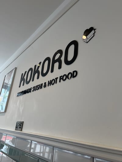 The image shows a ceiling with the words "KOKORO HANDMADE SUSHI & HOT FOOD" in black lettering. A small, stylized picture of what appears to be sushi is positioned above the word "KOKORO". An electrical outlet is near the tiles on the right side of the image. The image was taken in Eastbourne, United Kingdom.