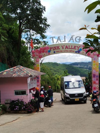 A silver van with its headlights on is parked on a road in front of a pink ticket booth. The van has a yellow license plate that reads "61B 030.58". Three people are standing near the ticket booth, and two people are on motorcycles behind the van. One of the people near the booth is wearing an orange helmet and appears to be handing something to another person. The ticket booth has a colorful, floral-patterned roof and signs with prices. Behind the booth and the van, a large, decorative archway with flowers spells out "TAJ AG PINK VALLEY THUNG LUNG HONG". The scene is set against a backdrop of lush green hills and a cloudy sky. The overall atmosphere suggests an entrance to a tourist attraction.