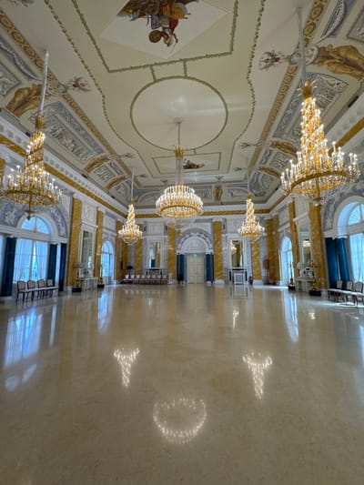 This is a wide-angle, eye-level photograph of a grand, empty ballroom in the Konstantinovsky Palace in Strelna, Russia. The room is exceptionally well-lit, with natural light streaming through large arched windows on the left side, and the bright illumination from multiple ornate, multi-tiered golden chandeliers. The highly polished, light-colored marble floor reflects the chandeliers and the architectural details of the room, creating a shimmering, ethereal effect.
The walls are a combination of white plaster, with intricate relief carvings, and columns clad in yellow and grey veined marble. Above the columns, the ceiling is painted with elaborate frescoes depicting mythological scenes and decorative borders in muted colors. The windows are tall and arched, draped with sheer white curtains. Several rows of chairs are visible against the walls in the distance, but the room is otherwise devoid of people.
The overall impression is one of opulence and spaciousness, with a focus on classical architectural grandeur and decorative artistry. The room appears immaculate and well-maintained, suggesting it is a significant venue for events.