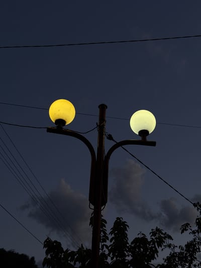The image shows a street light with two globe-shaped bulbs illuminating the night sky in Ogradna, Bulgaria. The sky is a dark blue, with scattered clouds partially obscuring the view. Power lines are visible stretching across the sky, and the silhouette of a tree is present on the right side of the frame. The street light is a dark color with a curved design. The upper bulb emits a pale yellow light, while the lower bulb appears more distinctly yellow.