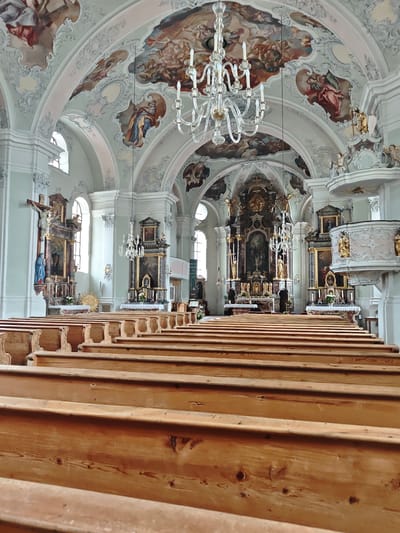 The image shows the interior of a church in Filzen, Austria. The perspective is from the pews looking towards the altar. The pews are made of light-colored wood and are arranged in rows. The walls and ceiling are ornately decorated with frescoes and stucco work. A large chandelier hangs from the ceiling. The altar area features an elaborate altarpiece, paintings, and sculptures. There are also religious figures visible on the walls and on the pulpit. The scene is well-lit, suggesting the presence of natural light from windows and artificial lighting. There are no people visible in the image.