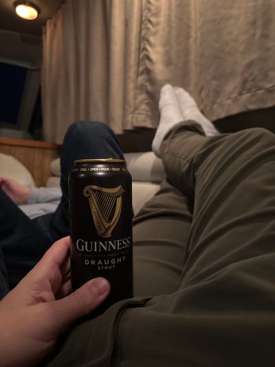 A person is holding a can of Guinness Stout in their right hand while seated. The can is black with gold lettering and the Guinness harp logo. The text on the can reads "GUINNESS", "ST JAMES'S GATE DUBLIN, IRELAND", and "DRAUGHT STOUT". The person is wearing olive green pants on top and blue jeans on the bottom. They also have white socks on. The background includes a curtained window and possibly a bed or seat.
