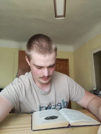 A young man with short blonde hair and a light brown t-shirt sits at a wooden table, engrossed in a book. He has a light mustache and stubble. The room is lit by natural light, suggesting it is daytime. The walls are painted yellow, and a wooden door is visible in the background. A light fixture hangs from the ceiling. The man's gaze is directed downwards at the open book, which appears to be an academic or historical text, with dense print and illustrations. The book has a dark purple marker or bookmark peeking out. The scene conveys a sense of quiet study or focused reading. The overall impression is that of a casual, indoor setting.