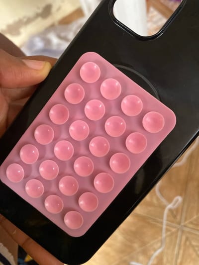 A close-up, high-angle shot shows a pink pop-it toy attached to the back of a black phone case, held by a person's left hand with visible fingernails. The pop-it toy has a grid of twenty-two pink, bubble-like protrusions arranged in five rows. The toy is placed diagonally on the phone case. The phone case has a cutout for a camera. The background consists of a wooden surface with a white string coiled on it, and a blurred white and beige patterned fabric. The scene is indoors, and there is no information to suggest it is in Tavo, Nigeria.