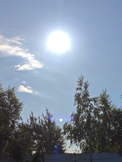 The image depicts a bright, sunny sky with the sun prominently positioned near the top center, emitting a strong glare. A few scattered white clouds are visible in the upper left quadrant. The lower half of the image is dominated by the silhouettes of several trees, their dark green foliage contrasting with the bright sky. In the very bottom of the frame, a muted grey fence runs horizontally. The scene appears to be an outdoor environment in Краснадворскі сельскі Савет, Belarus.
