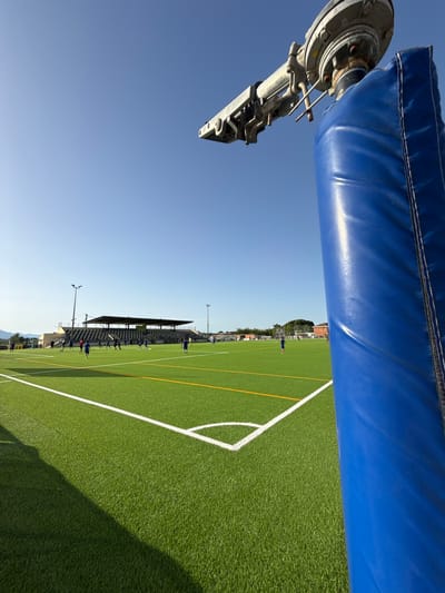 The image depicts an artificial turf soccer field in Vidreres, Spain. A blue padded post is in the foreground, partially obscuring the view of the field and a grandstand in the background. Several individuals, appearing to be soccer players, are scattered across the field, some engaged in activity. The sky is clear and blue, suggesting a sunny day. The scene is set in a sports facility with lighting poles and surrounding buildings visible.