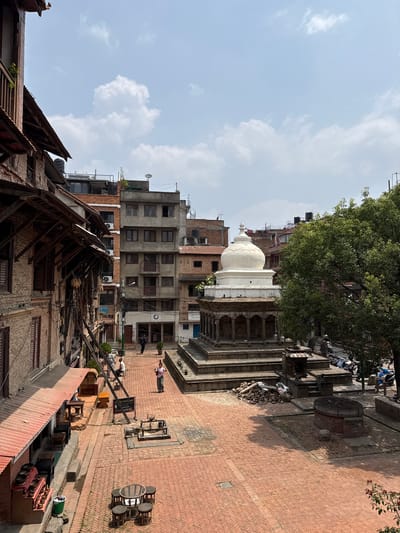 The image shows a street scene in Lalitpur, Nepal. A brick-paved street is lined with buildings of varying heights and architectural styles. Some buildings appear to be older with wooden beams and traditional Nepali architecture, while others are more modern concrete structures. A white stupa-like structure with a rounded dome is visible among the buildings. A large tree is partially visible. Several people are walking on the brick street. The sky is blue with scattered clouds. There are outdoor seating arrangements along with some old machinery and building material stored along the brick-paved street.