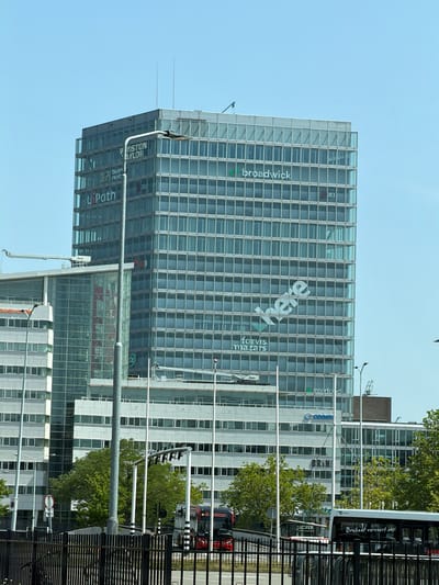 The image shows a contemporary glass-fronted office building in Eindhoven, Netherlands. The building's facade is covered in a grid of windows, reflecting the clear blue sky. Prominently displayed on the building are several company names, including "broadwick", "JLL", "U Path", "here", "forvis mazars", and "Interfood". A bus with text "Brabant vervoert ons" is visible in front of a neighboring building, suggesting public transportation is active in the area. Trees with lush green foliage are interspersed between the buildings, indicating a pleasant, possibly spring or summer day. The overall scene conveys an urban business environment.