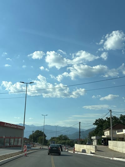 The image depicts a street scene in Radanovići, Montenegro, on a clear day with scattered white clouds. The foreground shows a paved road with yellow lane markings, bordered by construction materials and barriers on the right and a building with red trim and signage that reads "Auto Centar Tomicić" on the left. Several cars are visible on the road, with one black car in the immediate foreground. Streetlights are present along the road. In the background, trees and residential buildings are visible, with a range of mountains under a bright blue sky. Power lines crisscross the scene.