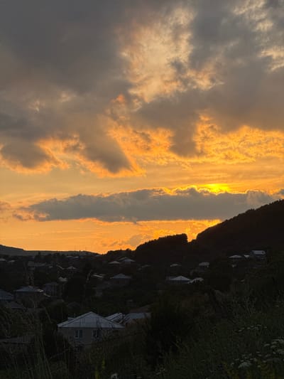 The image depicts a sunset over the town of Margahovit, Armenia. The sky is filled with dramatic clouds painted in hues of orange and yellow, with the sun partially obscured by a thick, dark cloud. Below the sky, a silhouette of hills covered in trees frames the landscape. In the foreground, the dark shapes of houses and trees are visible, suggesting a rural or semi-rural setting. Some houses have visible rooftops. The overall impression is of a serene, natural landscape at dusk.