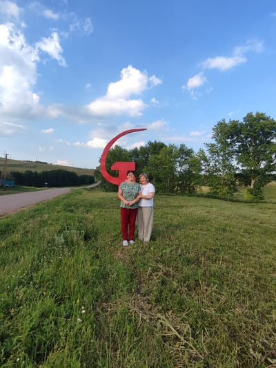 Two women stand in a grassy field in front of a large, red hammer and sickle symbol. The woman on the left wears a green and white patterned shirt and red pants. The woman on the right wears a white shirt and beige pants. Behind them, a road winds through a hilly landscape with trees and a blue sky with white clouds. The location is identified as Karabash, Russia.