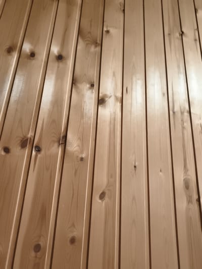 The image shows a close-up, angled view of a wooden wall paneled with vertical planks. The wood is light brown with visible grain patterns and knots. The planks have a tongue-and-groove or similar joinery, creating a linear texture. The lighting creates reflections and shadows on the surface, highlighting the three-dimensional quality of the paneling. The image does not provide any information about the city of Sputnik, Russia, or any people, actions, or specific environmental context beyond the wall itself.