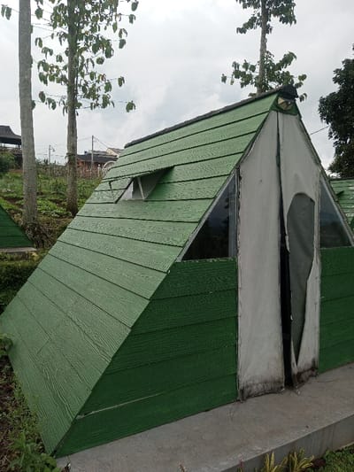 The image depicts an A-frame dwelling structure situated outdoors in Abbireddygudem, India. The structure features steeply angled walls and a roof clad in dark green, horizontal, wood-textured siding. The front facade presents a central opening covered by torn and weathered light grey fabric, flanked by two triangular clear glass panels. A small, triangular vent or window projects from the upper left side of the green roof. A circular decorative element or vent is visible at the peak of the front gable. The structure rests on a flat concrete foundation surrounded by sparse soil and low-growing vegetation. In the background, multiple tall, slender trees with green foliage rise against an overcast sky. Distant residential or communal buildings are visible on a higher, sloping terrain, crisscrossed by overhead utility lines. A second, similar green A-frame structure is partially visible to the lower left. No individuals or active interactions are present in the scene.