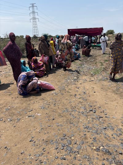 The image depicts an outdoor scene with a group of people, predominantly women wearing traditional clothing, gathered in a dusty, arid environment. In the background, a large electricity pylon stands tall against a clear sky, suggesting proximity to infrastructure. A maroon-colored canopy or tent structure is visible further back, possibly indicating a gathering point or shelter. Several individuals are seated or standing in groups, with some appearing to be engaged in conversation or awaiting something. The ground is covered in dirt and scattered stones. The overall atmosphere suggests a humanitarian or community gathering in a rural or semi-urban setting within Ad-Damazin, Sudan.