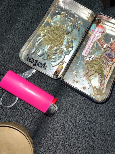 A slightly elevated, angled shot shows an open metal container resting on a dark, textured surface. The container, divided into two sections, holds what appears to be loose cannabis. One section also contains a partially smoked rolled cigarette. Writing is visible on the side of the container. Adjacent to the container is a pink lighter and a pair of scissors. Part of a circular metal object is visible in the lower right corner. The photo was taken in Llavallol, Argentina.