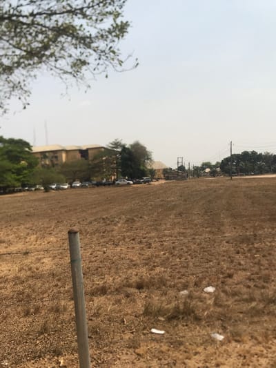 The image, taken in Umudike, Nigeria, captures an expansive outdoor landscape under a bright, hazy sky. The foreground and much of the midground are dominated by a large field of dry, light brown vegetation, suggesting a period of drought or a dry season. Scattered pieces of white debris are visible on the parched ground in the foreground. A grey metallic pole stands vertically on the right side of the frame, partially obscuring the field.
In the distance, along the left side of the horizon, a cluster of light-colored buildings is visible, interspersed with trees of varying foliage density. A line of cars, mostly light-colored, are parked along what appears to be a road or path leading towards these structures. Utility poles and power lines are also visible in the background. A tree with delicate, lighter green branches is partially cropped into the far left foreground.
The sky is a uniform, bright white, indicating daytime, possibly with atmospheric haze common during dry seasons or the Harmattan. No specific activities or events are discernible, and no people are visible in the scene, which conveys a sense of stillness. No visible text is present.