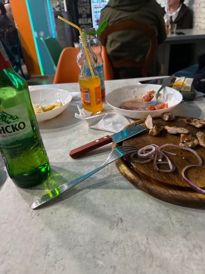The scene depicts a table within an indoor dining establishment in Gorna Mitropolia, Bulgaria. The table surface is light grey, possibly concrete or laminate. In the foreground, a partially consumed green glass bottle of "Pirinско" beer stands upright. To its right, a white bowl contains yellow food remnants, possibly fries. A clear glass bottle of orange soda (Fanta) with a yellow plastic straw is positioned next to a crumpled white paper napkin. Beyond this, a second white bowl contains a small amount of residual salad, including red tomato pieces, with a silver fork resting within it. To the right of the napkin, a wooden cutting board holds several pieces of cooked meat, likely poultry or pork, alongside several thin slices of red onion. A knife with a brown wooden handle and a silver blade lies on the board next to a separate silver fork. In the blurred background, a person wearing a dark green jacket is seated with their back to the viewer, facing a table. An orange chair is visible between the table in the foreground and the background figures. Vertical illuminated strips, one orange and one teal, are visible on a wall. Further back, a sign with "WINE BEER" text is partially visible above what appears to be a refrigerated display.