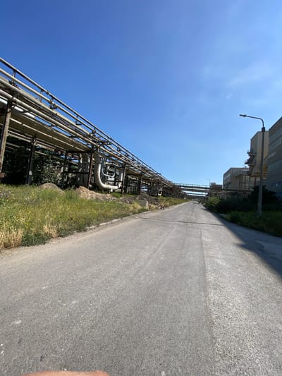 This media file depicts an outdoor industrial setting in Poland, likely during daytime on a clear, sunny day. The primary subjects are a vast network of industrial pipes and infrastructure, characteristic of a chemical plant or similar facility. These pipes, some appearing aged and metallic, are elevated on a metal framework that runs alongside an asphalt road.

The scene is dominated by the industrial structures and the road, with a strip of vegetation and tall grass bordering the road and the pipes. In the background, modern industrial buildings are visible, suggesting an active or recently operational site. A street lamp on a pole is also present, further indicating an organized, if somewhat stark, environment.

There is no visible activity or event taking place; the image appears to be a still shot capturing the infrastructure of the location. The clear blue sky suggests good weather conditions. The lack of people or any signs of recent activity gives the image a sense of quiet or dormancy. The visual composition, with the road receding into the distance and the industrial elements stretching along one side, creates a perspective that emphasizes the scale and complexity of the industrial site. No text is visible in the image.