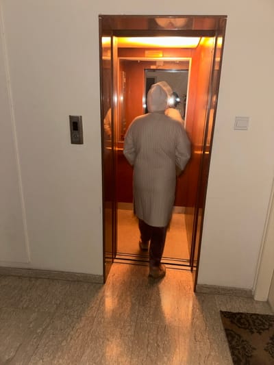 The image, correctly oriented, shows a person stepping into an elevator within an indoor hallway. The subject, viewed from behind, is wearing a light-colored, possibly striped, long-sleeved top or jacket, dark trousers, and light-colored footwear, along with a light-colored head covering. Significant motion blur is visible on the person and parts of the elevator's interior, indicating movement during the photograph.

The hallway features white walls and a light-toned, possibly marble or tiled floor, with a section of a patterned rug visible on the far right. The open elevator doors reveal an interior bathed in a warm, orange glow, likely from internal lighting, which reflects off polished surfaces. The elevator's frame appears to be a metallic, possibly bronze-toned, material.

On the white wall to the left of the elevator entrance, a silver-colored elevator call panel is mounted, featuring a small, unreadable digital display and buttons. Above and slightly to the left of the elevator, a plain white light switch is present on the wall. The warm lighting from the elevator and an unseen source casts soft shadows on the hallway floor. No discernible text is clearly readable in the image. The scene suggests an everyday moment of transit within a modern building, situated in Egkomi Lefkosias, Cyprus.