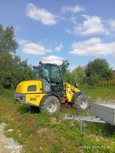 This is a photograph of a yellow Wacker Neuson WL48 wheel loader parked on a grassy area with a small utility trailer attached. The wheel loader is in the foreground, angled towards the left side of the frame, with its front bucket raised. The cab of the loader has tinted windows and a single orange warning light on top. The wheel loader has "WACKER NEUSON" branding and model number "WL48" visible on its body. There are red flowers growing around the front tire of the loader.

The background consists of lush green trees and bushes, with a few tall cypress trees on the right side of the frame, suggesting a rural or suburban setting. The sky is blue with scattered white clouds.

A silver utility trailer is parked behind the wheel loader, connected to its hitch. The trailer appears to be empty. In the lower left corner of the image, there is text that reads "POCO C75". In the lower right corner, there is a date and time stamp: "08.06.2026 09:56".

The overall impression is a sunny day, possibly in a construction or landscaping site, given the presence of the heavy machinery and trailer. The location is identified as Kiełczów, Poland.