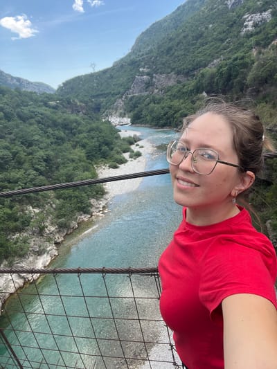 A young woman in a red t-shirt smiles at the camera, wearing glasses and earrings, in an outdoor setting on a bridge or walkway with metal cables and wire mesh. Below her is a turquoise river bordered by rocks and dense green trees covering the steep mountain terrain. The sky is light blue with a few clouds visible in the background. The location is inferred to be in the city of Unknown, Montenegro.