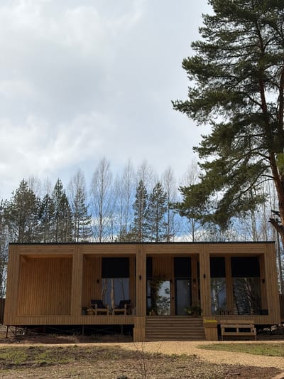 A modern, two-story cabin made of light-colored horizontal wooden planks stands in a tranquil, forested setting in Mishkino, Russia. The building, positioned on the right side of the frame, features multiple large, dark-paned windows that reflect the surrounding natural environment. A wooden deck or porch area extends from the front, furnished with dark outdoor chairs and a potted plant, suggesting an inviting outdoor living space.

Dense, dark green pine branches with needles hang prominently over the upper left portion of the image, contributing to the woodland atmosphere. Beyond the immediate foreground, a mixed forest landscape unfolds, characterized by numerous bare deciduous trees alongside more evergreen pines, suggesting a transitional season such as early spring or late autumn. The sky, visible through the trees, is partly cloudy with patches of clear blue. The ground around the cabin is covered with dry earth and sparse vegetation.

No people are visible in the scene, which evokes a sense of peace and natural seclusion. The lighting indicates it is daytime. There is no discernible text within the image. The overall impression is of a quiet, modern retreat seamlessly integrated into a rustic, natural environment.