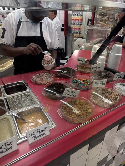 An employee prepares an ice cream cup at an ice cream shop in Boca Raton, United States. The employee, wearing a white shirt, black apron, and grey hat, holds an ice cream cup filled with pink ice cream and sprinkles. Above the employee, there's a display case filled with ice cream toppings like coconut flakes, Heath Bar pieces, M&Ms, butterfinger pieces, chocolate chips, and sprinkles. Below the employee are various syrups and toppings, including hot peanut butter. Signs label each topping and syrup. In the background, there are ice cream cones and more cups for customers. The tiles on the side wall are black and white. A price tag is seen at the bottom left of the image.