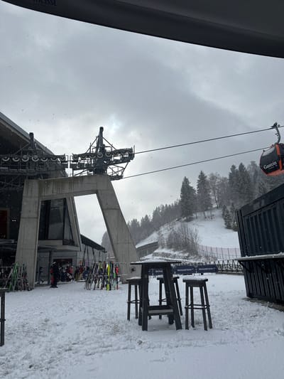 The image depicts an outdoor scene at a ski resort during winter, identified as the Gastein ski region, specifically in Bad Hofgastein, Austria, based on visible signage. The environment is characterized by a significant accumulation of snow covering the ground and the surrounding mountainous terrain, with evergreen trees on the hillsides. Light snowfall is visible against an overcast, grey sky.
In the foreground, a dark-colored outdoor bar table with three matching stools stands on the snow-covered ground; a second similar table and stool are partially visible to the left. All surfaces of these objects are dusted with snow. To the immediate right, part of a dark, corrugated structure, possibly a kiosk or container, is visible, also with snow on its horizontal surfaces.
The central and left midground are dominated by a large, modern concrete structure, which serves as a support pillar and terminal for a gondola lift system. This structure features a prominent A-frame-like base and supports a complex metal mechanism of pulleys and wheels at its apex, where gondola cables pass through. Behind this concrete structure, a contemporary building with dark-framed windows, likely the lift station or an associated facility, is partially visible.
In front of the building and near the concrete pillar, numerous pairs of skis and a few snowboards are organized upright in racks on the snow. Several individuals, dressed in winter ski attire, are present in this area, appearing to be standing or moving near the lift entrance or ski storage.
Running from the lift station towards the right, gondola cables are strung across the sky. An orange gondola cabin with "Gastein" written in white is visible on the right side of the frame, suspended from these cables. Below the gondola cables in the midground, a black banner or fence with "SKIGASTEIN" in white lettering is visible, extending along the base of the snowy slope. The hillside beyond is covered in snow and trees, with a distinct ski slope visible.